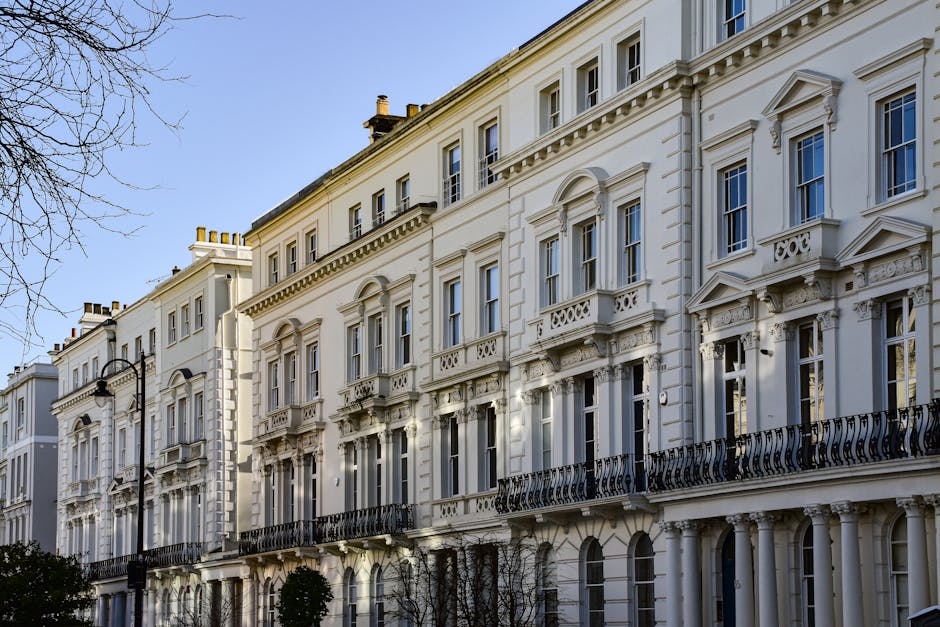 A row of elegant Victorian-style white terraced townhouses with ornate architectural details, including decorative mouldings, balustrades, and window pediments, set against a clear blue sky during daytime. The buildings feature multiple large sash windows with white frames, some with small balconies with intricate black wrought iron railings on the second floor. The façade appears freshly painted, with a smooth finish and subtle shadowing highlighting the architectural embellishments. In the foreground, there are leafless tree branches and a traditional streetlamp mounted on a black pole, suggesting an urban residential area. The ground level shows a clean pavement with small, neatly trimmed bushes positioned along the street. The scene is well-lit by natural light, emphasizing the historic character and clean condition of the buildings. This setting exemplifies a high-quality residential environment where private property maintenance and potential on-site rubbish clearance, such as by Rubbish Clearance Soho, may be relevant for property management or renovation projects.