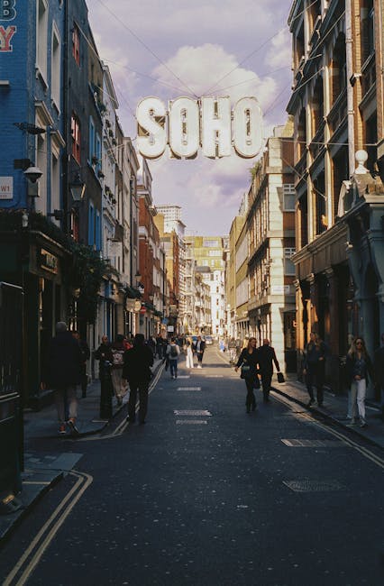 A busy urban street scene in Soho, London, with pedestrians walking on the pavement and crossing the road among classic Victorian-style buildings painted in various shades of blue, beige, and brick. The sky above is partly cloudy with a hint of blue, and a large illuminated sign spelling 'SOHO' hangs overhead, suspended between the buildings. The street is lined with small shops, cafes, and businesses, some with outdoor signage and awnings. The scene is well-lit with natural daylight, highlighting the textures of the building facades and the clothing of the pedestrians. This image captures the lively atmosphere of a central London district, illustrating an area where independent commercial activities and private disposal practices are common, reflecting the local environment thatRM rubbish clearance services, like Rubbish Clearance Soho, often serve for efficient waste removal in busy city streets.