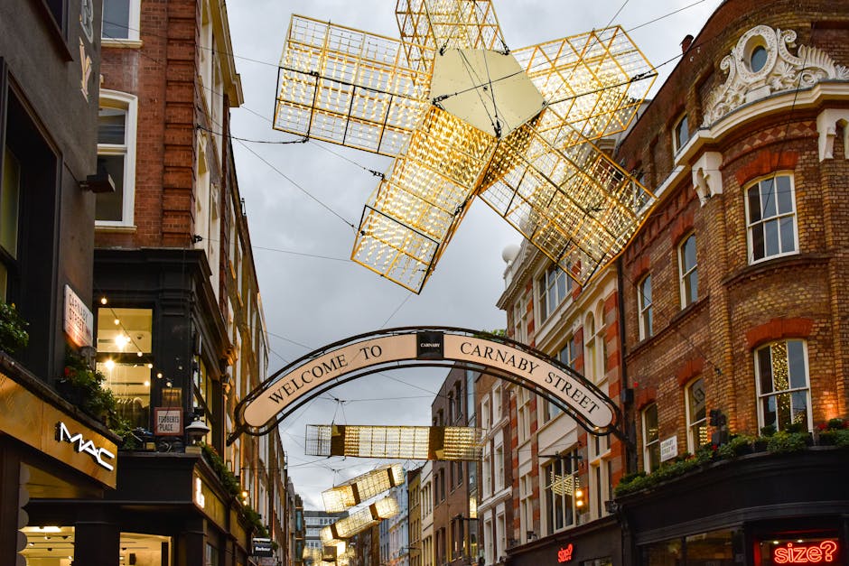 A narrow city street scene along Carnaby Street during daytime, featuring a large, modern geometric installation hanging overhead composed of illuminated golden cube-like structures made of metal frames. The installation is suspended by thin wires stretching between the surrounding brick and stone buildings, which display a mix of architectural styles, including ornate decorative elements and large sash windows. An arched sign reading 'Welcome to Carnaby Street' spans the entrance, positioned below the hanging installation. The façades of the buildings host various shopfronts, including a visible MAC cosmetics store with illuminated signage on the left, and other retail outlets with illuminated signs and window displays. Potted plants and window boxes adorn some of the buildings, adding greenery to the urban environment. Brightly lit shop windows contrast with the overcast sky, creating a lively shopping atmosphere. The scene captures an area known for independent and high-street retail, with the decorative lighting and architectural details supporting the local commercial character and alternative retail ambiance typical of the district.
