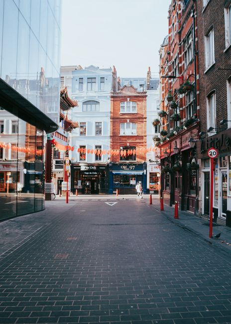 An urban street scene in Soho with a pedestrian walkway paved in dark grey bricks, flanked by multi-storey buildings on both sides, featuring a mix of red brick and light-colored facades. To the left, a modern glass-fronted building reflects the surrounding structures. On the right, traditional brick buildings with small shopfronts and potted plants visible at window level. Overhead, strings of orange lantern decorations stretch across the street, adding a decorative element. In the background, a white building with multiple windows and ornamental details rises behind the storefronts. Small red bollards line the edge of the pavement, separating pedestrian space from the roadway, which displays a 20 mph speed limit sign. The scene appears to be during daylight hours with natural light illuminating the area, and no vehicles or significant rubbish visible, exemplifying an orderly urban environment that could benefit from private waste management or rubbish clearance services by companies like Rubbish Clearance Soho, supporting alternative waste handling in busy city districts.