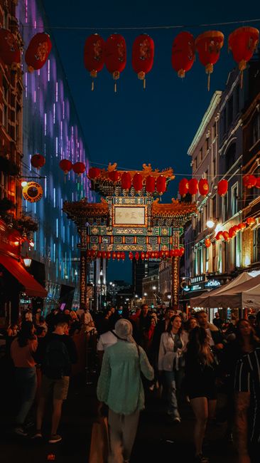A narrow city street scene along Carnaby Street during daytime, featuring a large, modern geometric installation hanging overhead composed of illuminated golden cube-like structures made of metal frames. The installation is suspended by thin wires stretching between the surrounding brick and stone buildings, which display a mix of architectural styles, including ornate decorative elements and large sash windows. An arched sign reading 'Welcome to Carnaby Street' spans the entrance, positioned below the hanging installation. The façades of the buildings host various shopfronts, including a visible MAC cosmetics store with illuminated signage on the left, and other retail outlets with illuminated signs and window displays. Potted plants and window boxes adorn some of the buildings, adding greenery to the urban environment. Brightly lit shop windows contrast with the overcast sky, creating a lively shopping atmosphere. The scene captures an area known for independent and high-street retail, with the decorative lighting and architectural details supporting the local commercial character and alternative retail ambiance typical of the district.