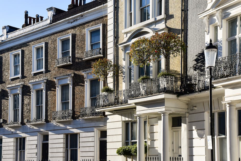 The image depicts a row of multi-storey residential buildings with Victorian architectural features, including sash windows and decorative wrought-iron balconies. The buildings are constructed with brickwork, some painted white and others displaying exposed brown bricks, with textured surfaces and contrasting finishes. In the foreground, there are two small trees with rounded foliage, planted in flower pots on the balconies, adding a touch of greenery to the urban scene. A traditional street lamp is positioned on the right side, casting a shadow on the white facade of one of the buildings. The scene is illuminated by daylight, with clear shadows indicating a sunny day. The presence of the buildings, along with the small trees on the balconies and the street lamp, suggests a typical residential street where private refuse collection and on-site clearance services, such as those offered by Rubbish Clearance Soho, may be relevant for managing rubbish and waste that accumulates in such environments, aligning with the context of alternative waste handling in densely built urban areas.